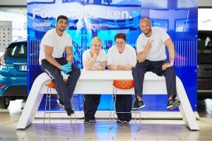 Vier Kolleg*innen sitzen und stehen an einem Tisch und lächeln in die Kamera.